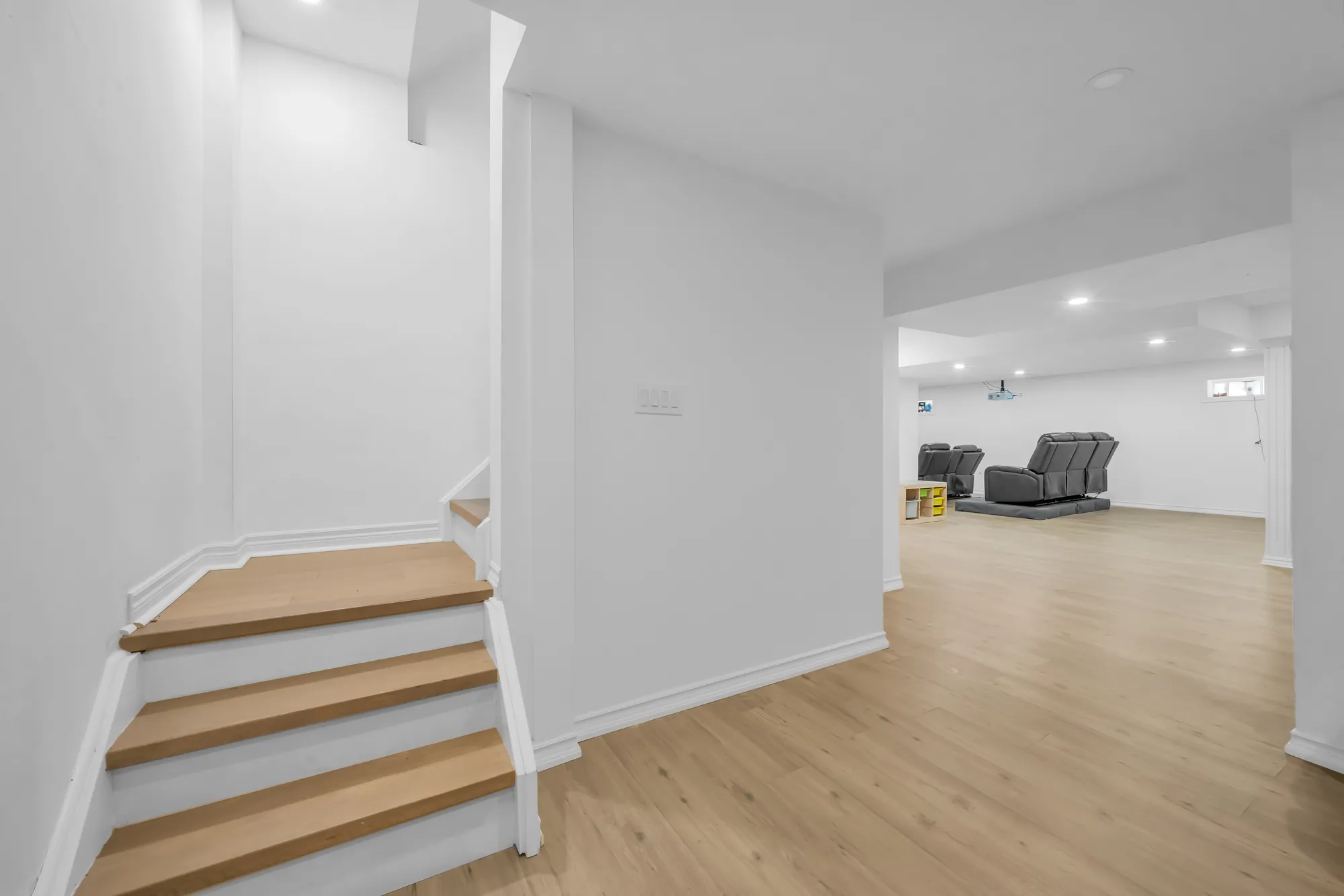 The foot of the basement stair, the open basement beyond: white walls, pot lights, light wood-look floors, two recliners