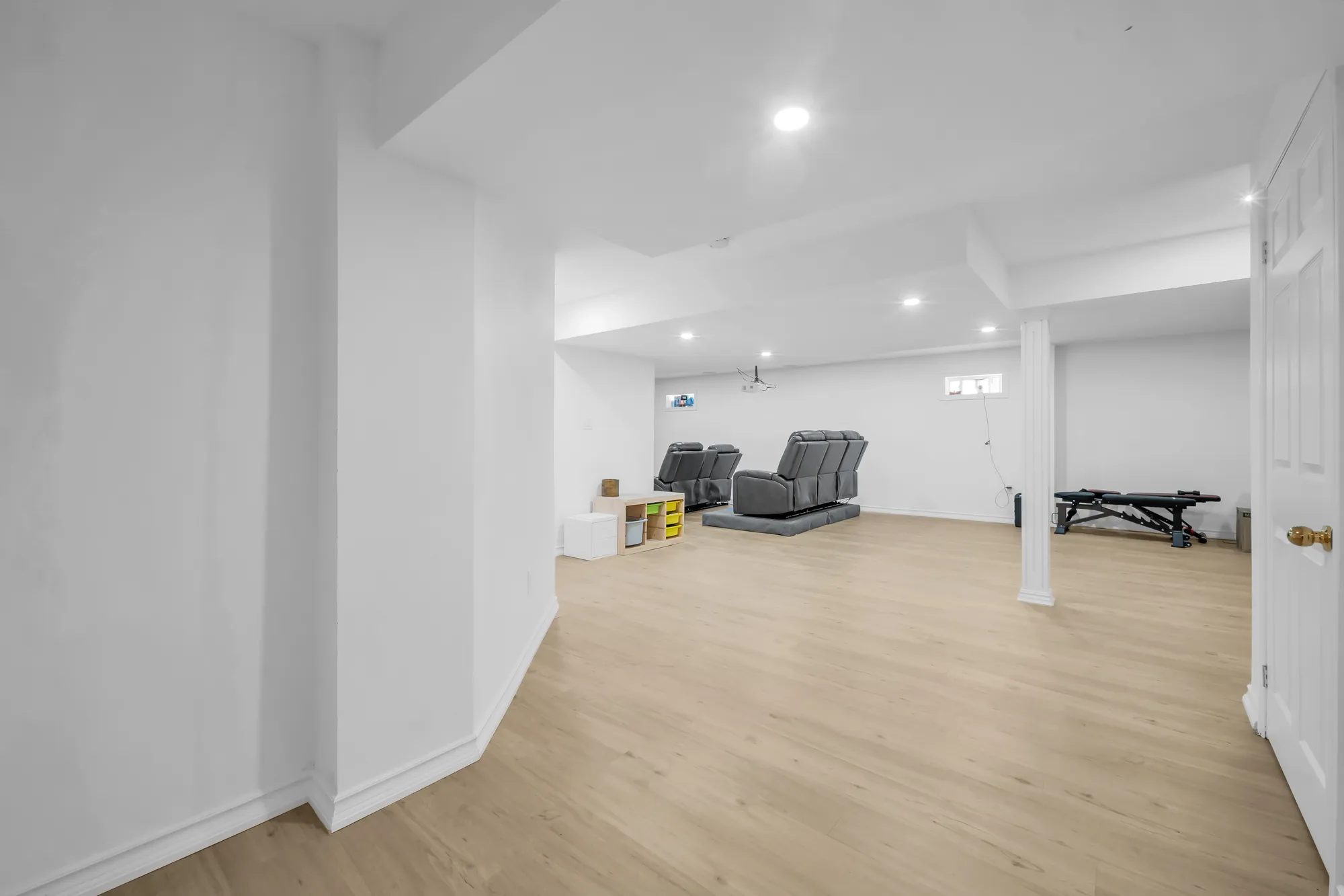 The open basement: white walls, pot lights, two recliners, cube shelves, a column, a door at the right