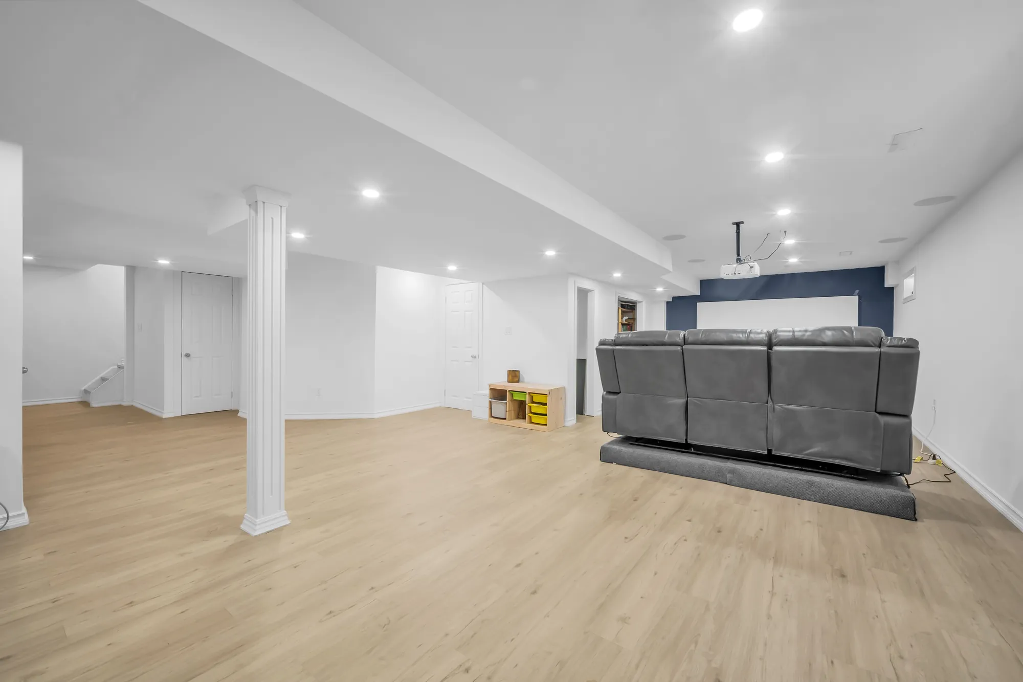 The basement toward a navy wall with a screen, a ceiling projector, a grey recliner sofa, doors along the far wall