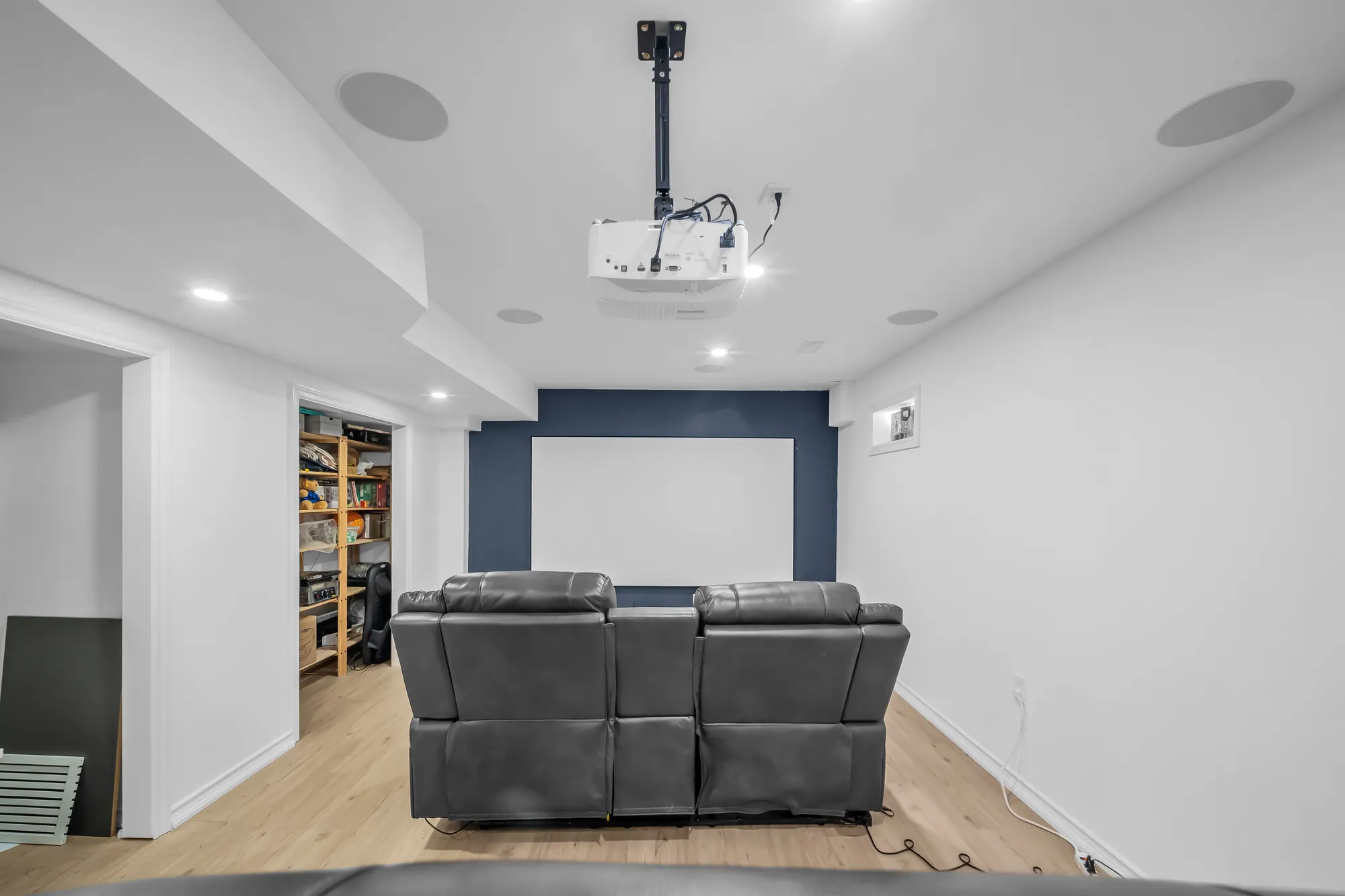 The projection wall: a screen on a navy wall, a ceiling-mounted projector, speakers in the ceiling, a recliner sofa, shelving through a door at the left