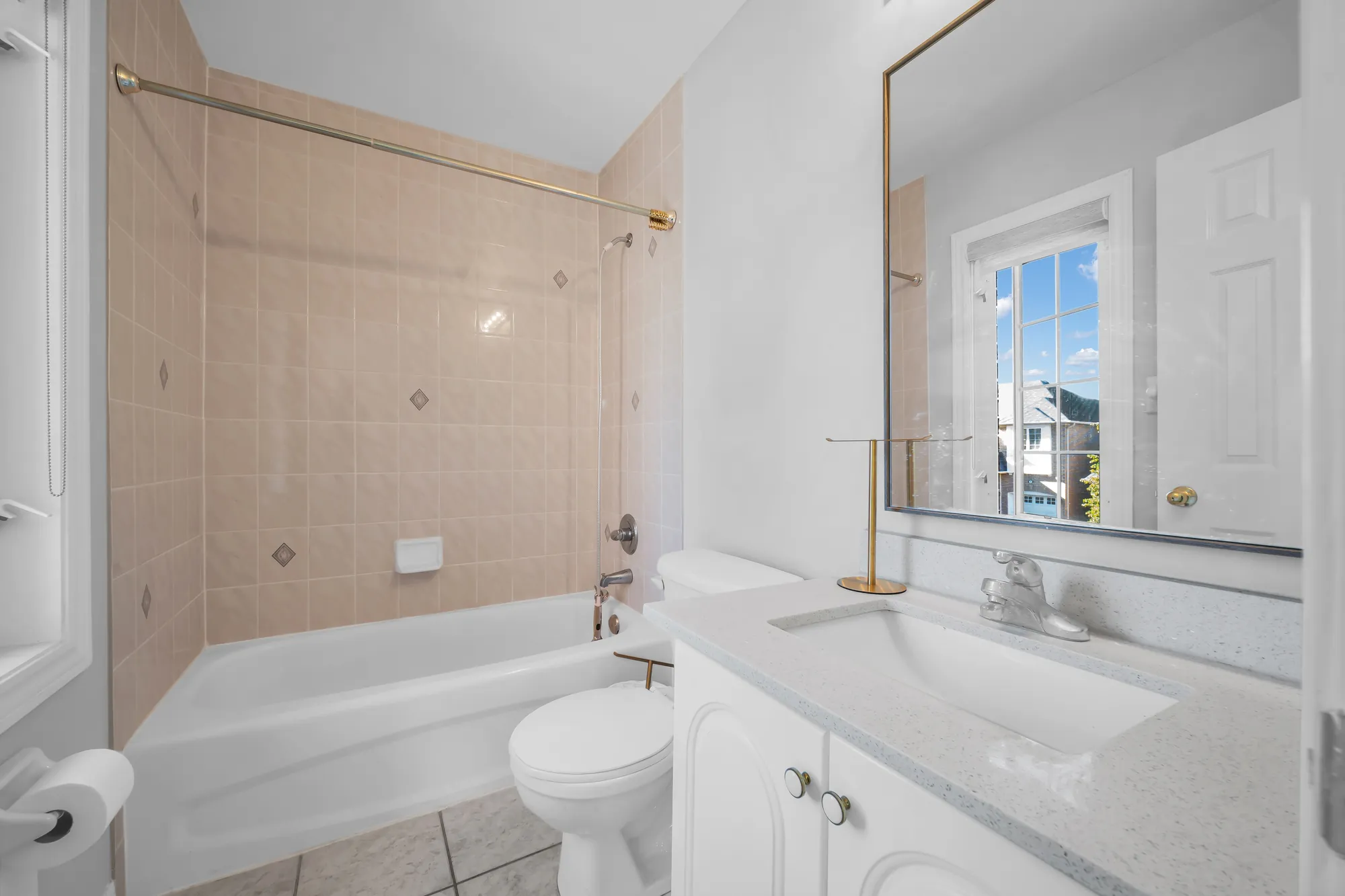 A bathroom: a tub with a pale pink tiled surround and a brass rail, a toilet, a white vanity, a brass-framed mirror, a window