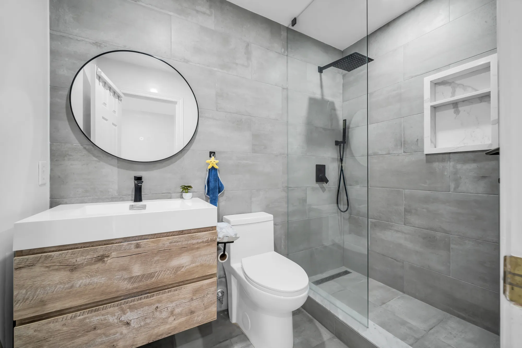 A bathroom off the upper hall: a glass shower with a black rain head, large grey tiles, a floating wood vanity, a round black mirror