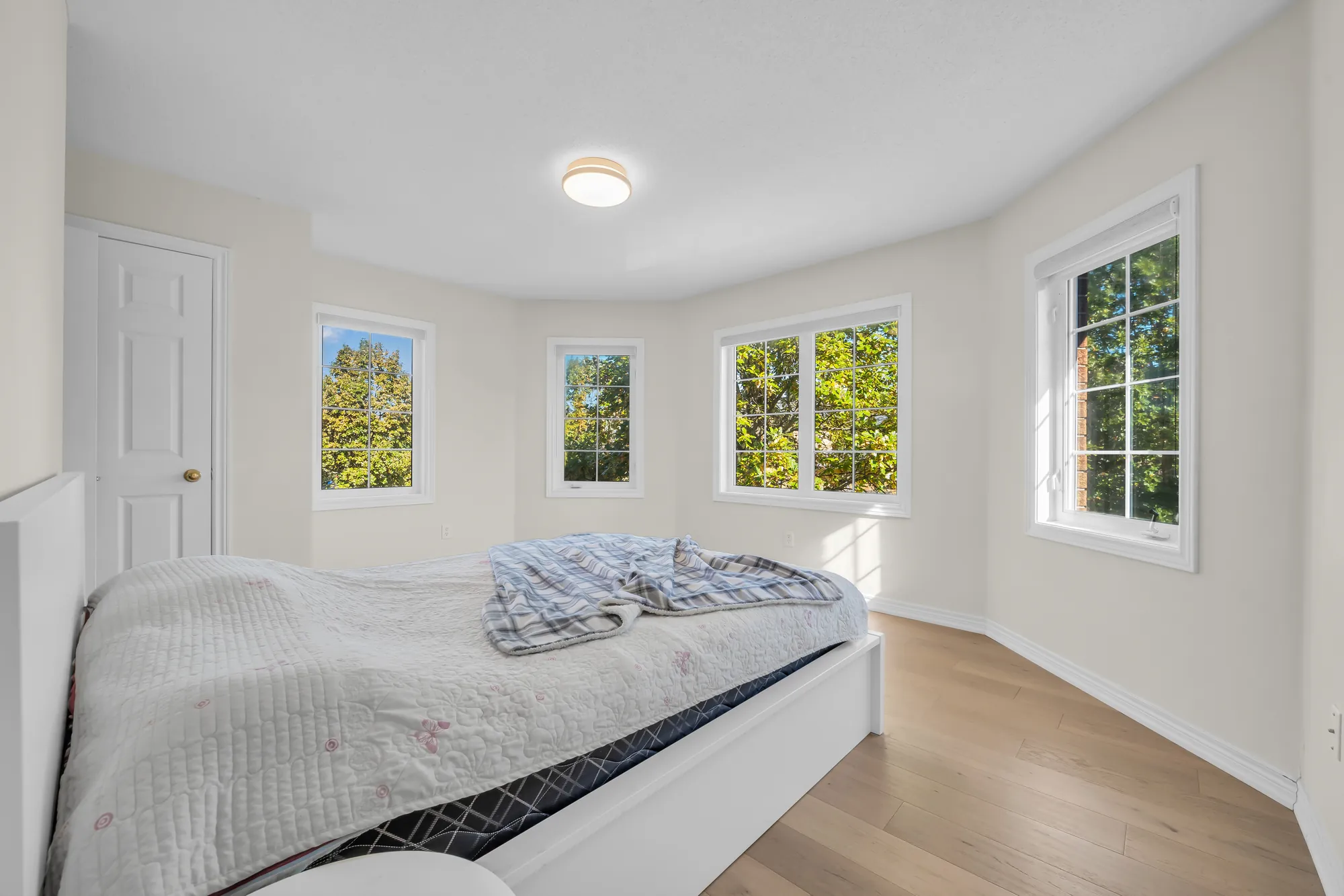 A bedroom with windows on three walls of a bay, a white bed, wood floors, a door at the left