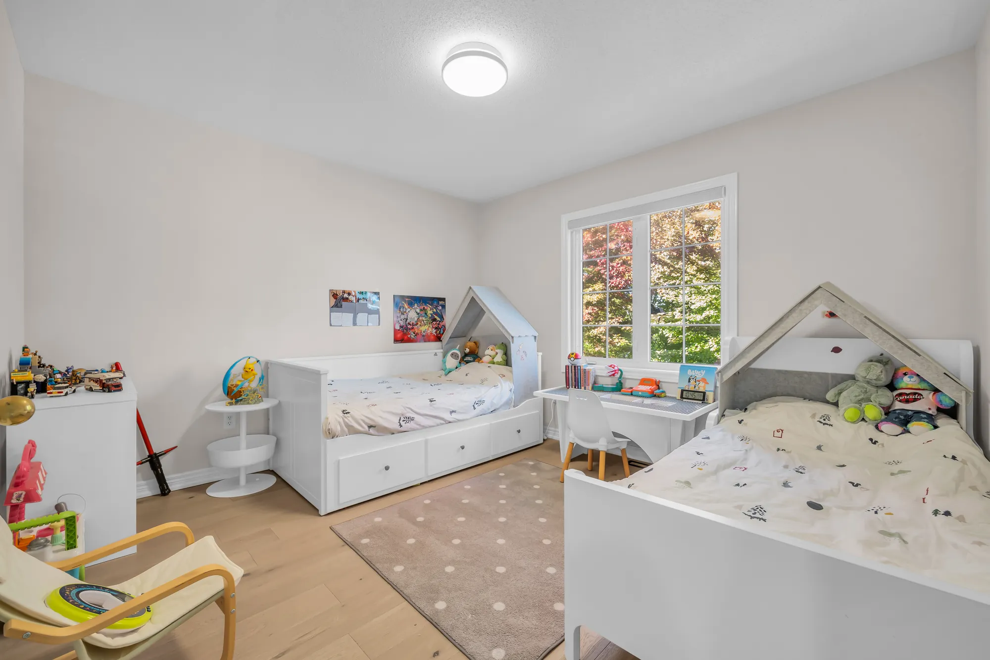 A child&rsquo;s bedroom: two white beds, one with a house-shaped frame, a small desk, a rug, toys on the shelves, a window onto trees