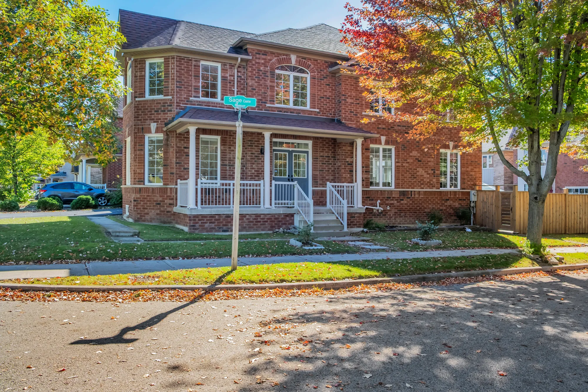 The house from the corner of the street: red brick on two storeys, a covered porch with white railings and steps up to the front door, a fenced yard at the right, a street sign and a maple at the corner