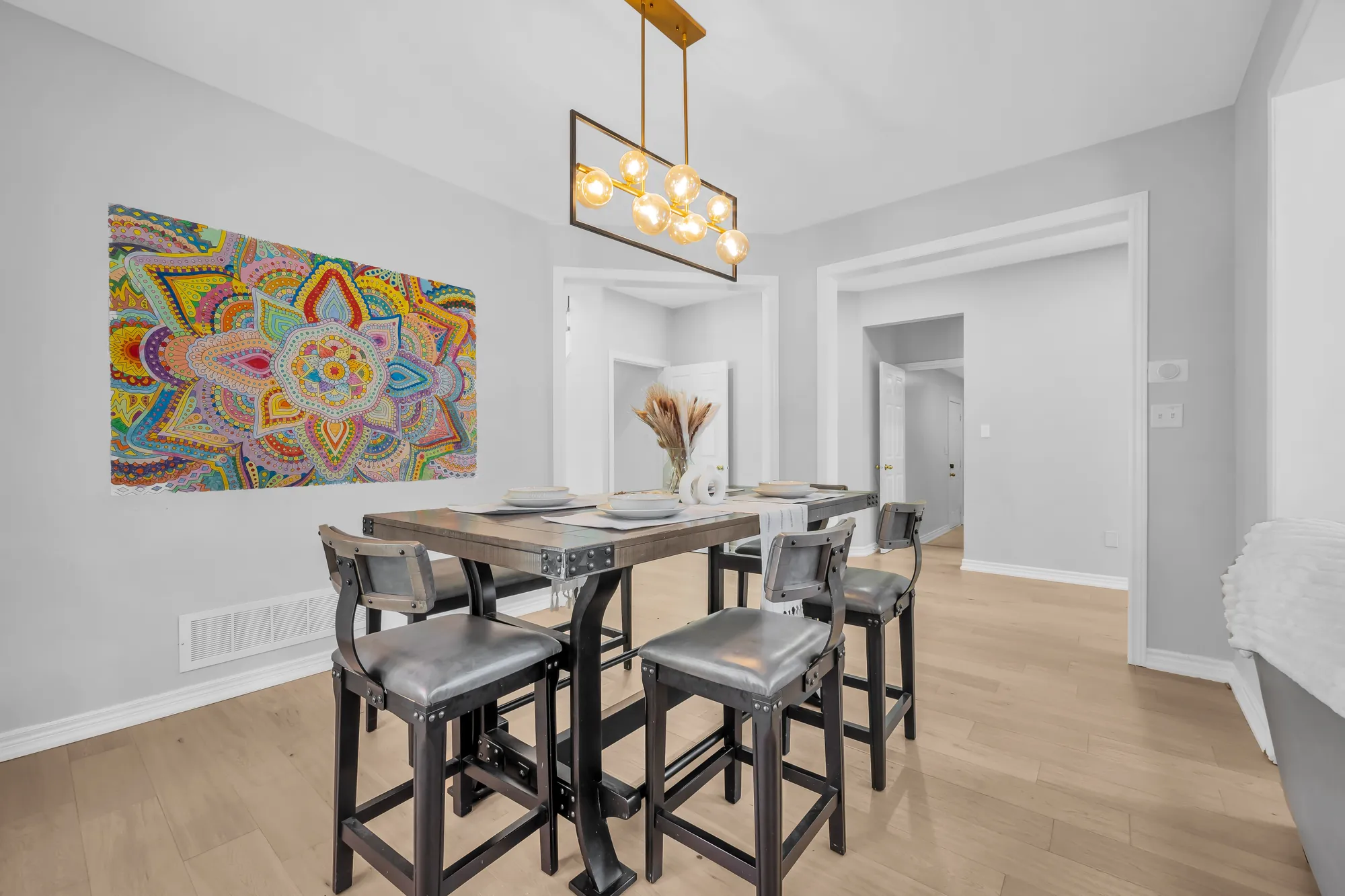 The dining room: a tall wood table with grey upholstered stools and a bench, a gold linear chandelier, a large colourful painting, windows onto the yard