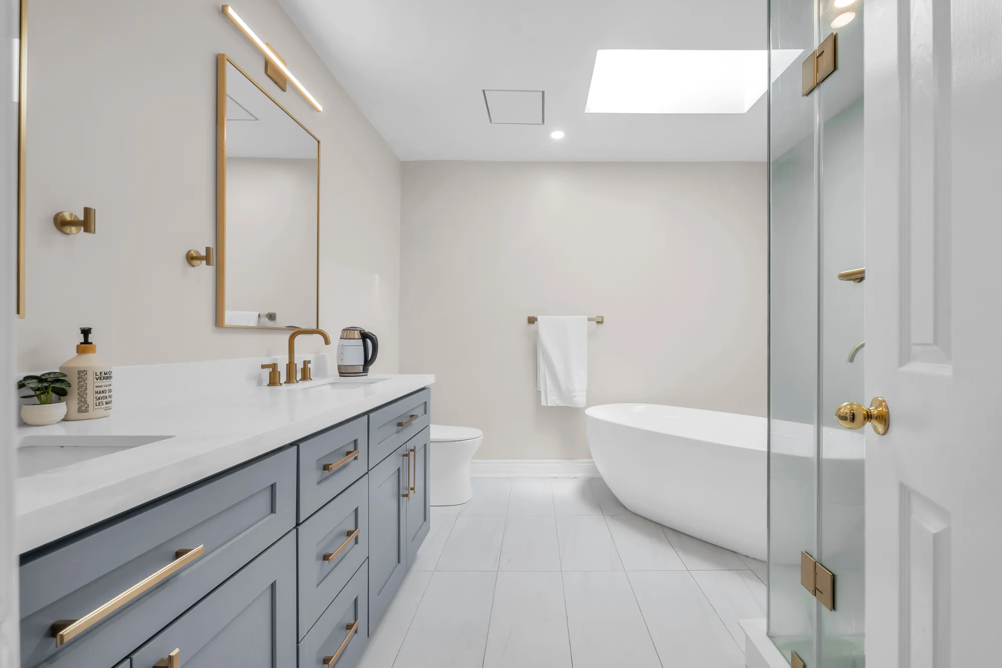 The primary bathroom: a double vanity in grey with brass fittings, a freestanding tub under a skylight, a glass shower