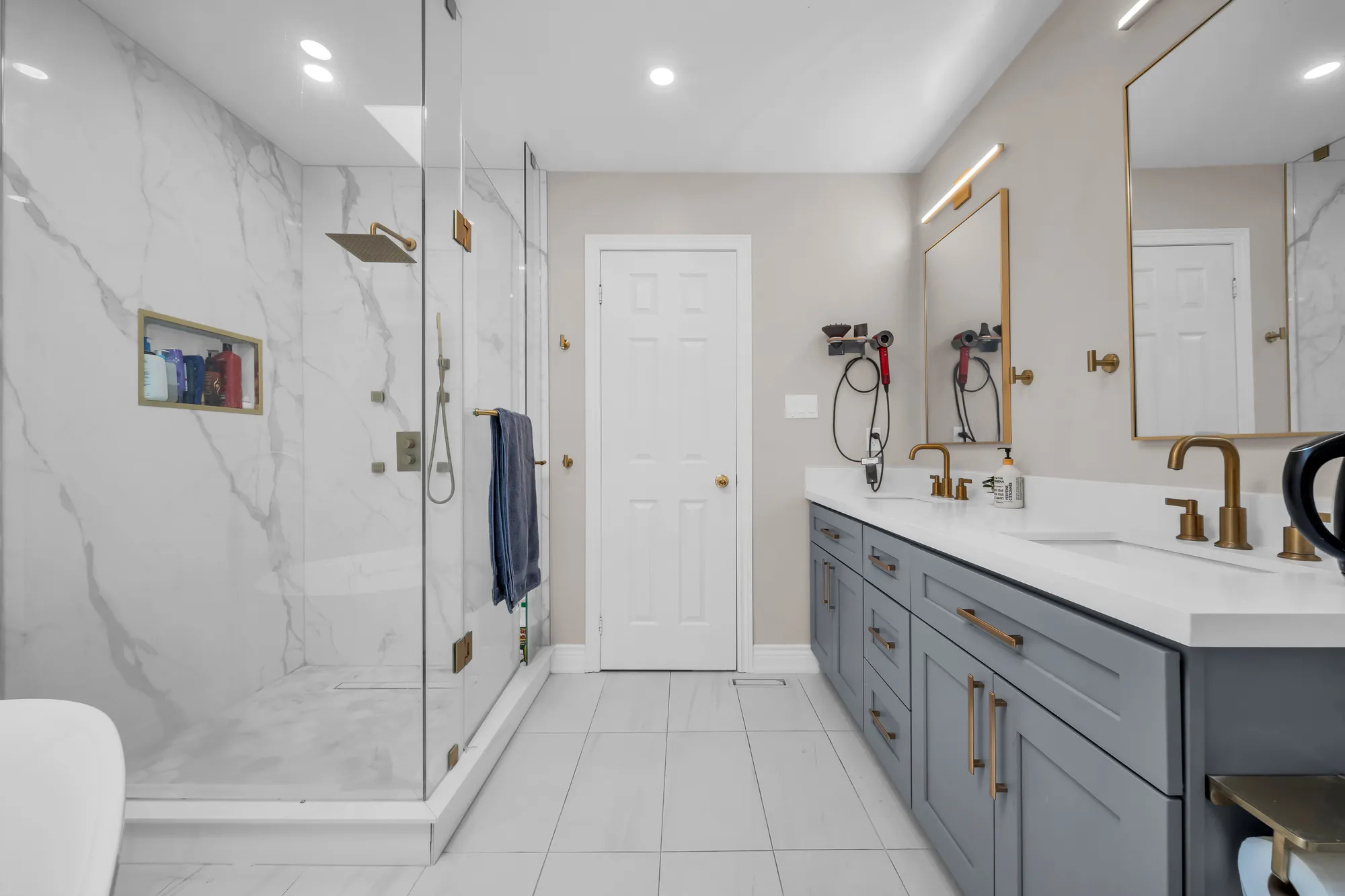 The glass shower lined in white marble-look tile with a niche, the grey double vanity and a gold-framed mirror, a door at the far end