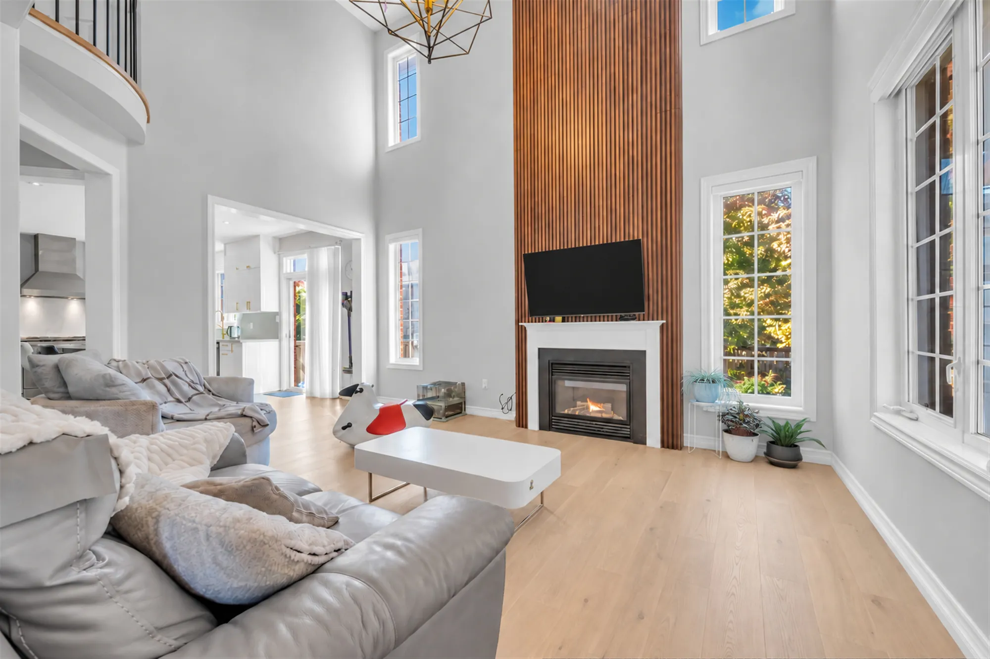The family room, two storeys high: a gas fireplace in a white mantel under a wall of vertical wood slats to the ceiling, a television, tall windows, grey leather sofas, a white coffee table