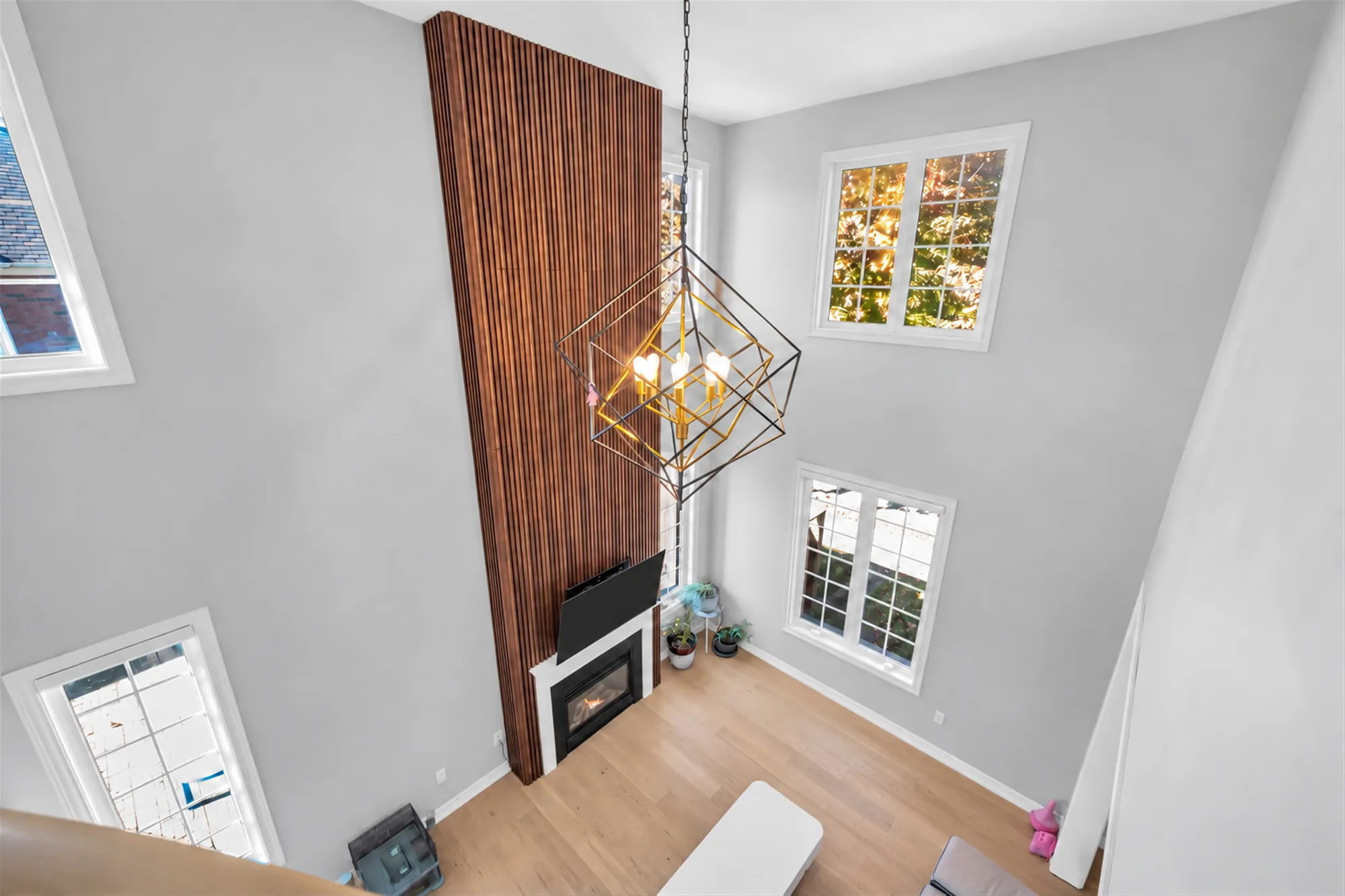 Looking down into the family room from the upper hall: the wood-slat wall rising to the ceiling, a geometric gold chandelier, the fireplace, two tiers of windows
