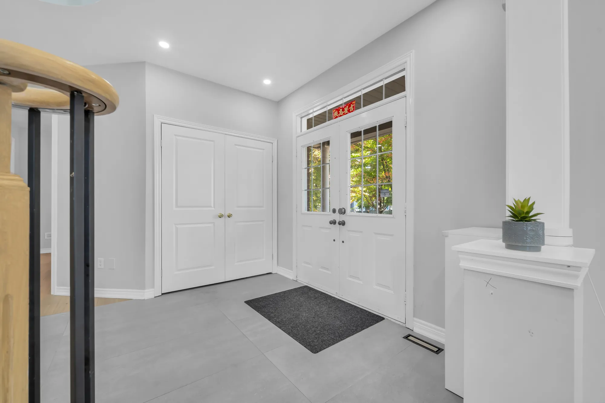 The foyer: a pair of front doors with glass panels and a transom, a double closet, a large grey tile floor, a mat, a white pedestal with a plant