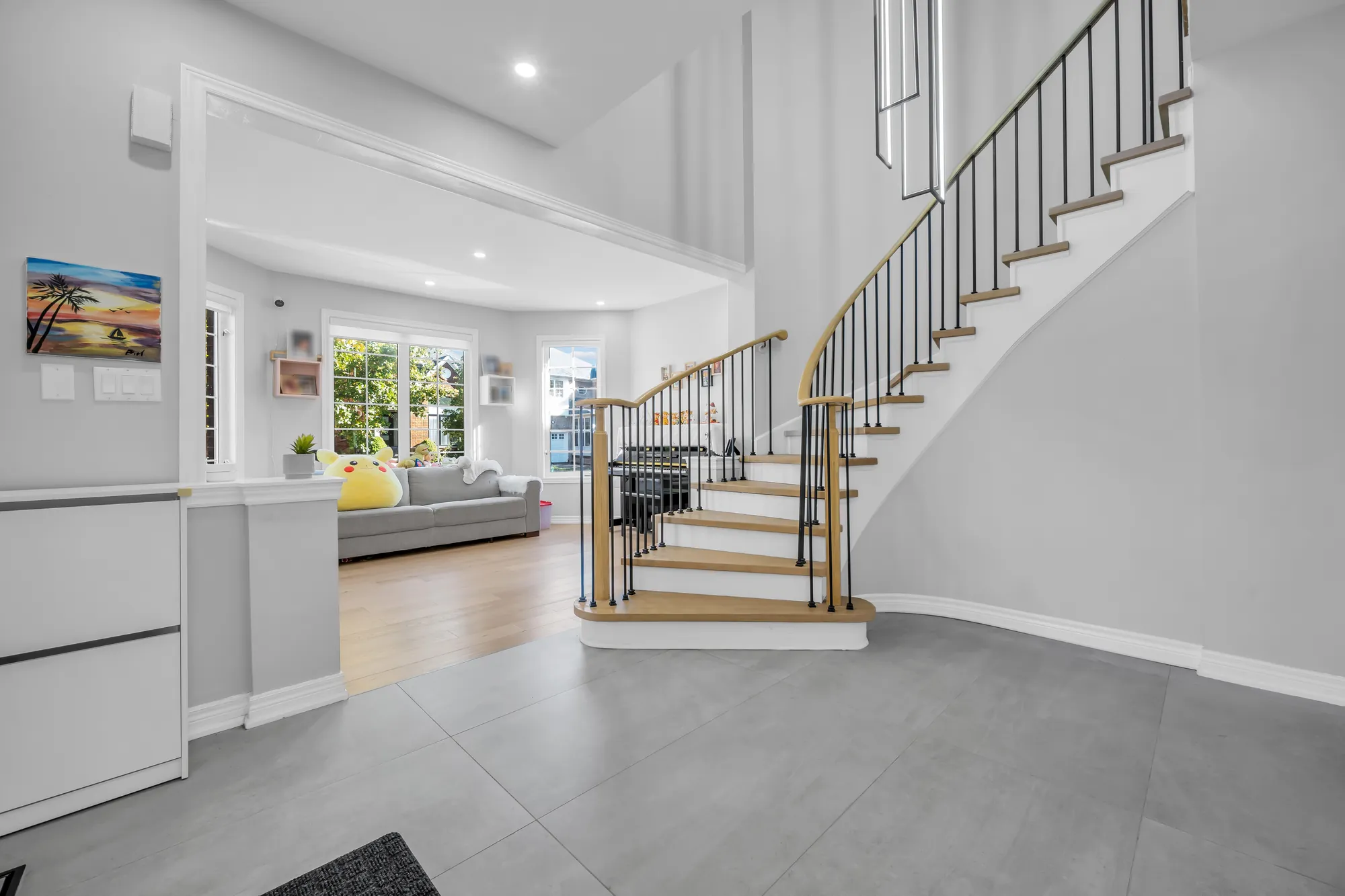 The foyer toward the curved staircase with an oak handrail and black spindles, the living room beyond with a grey sofa and windows onto the street