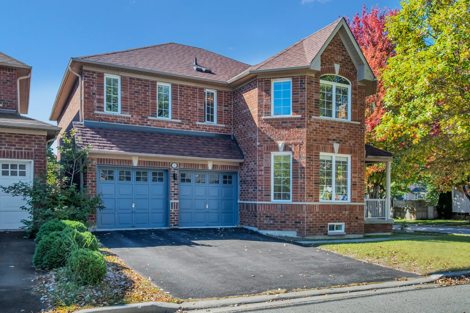 The front of the house by day: two storeys of red brick under a shingled roof, an attached double garage with two blue doors, a paved drive, a clipped hedge, maples in autumn colour at the right