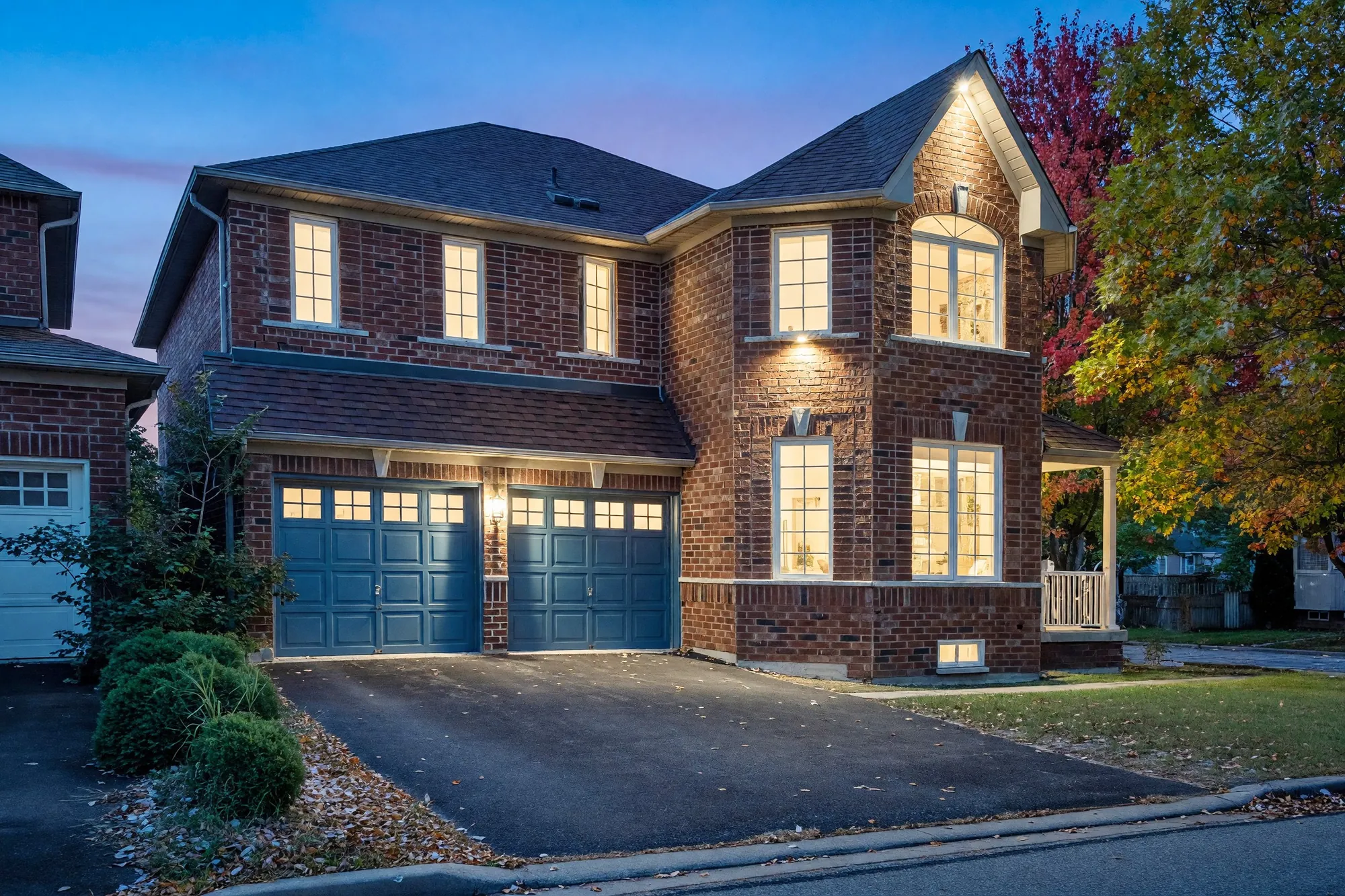 The front of 30 Pinecrest Street at dusk, an edited photograph: two storeys of red brick, an attached double garage with two blue doors, the windows lit, maples in autumn colour at the right