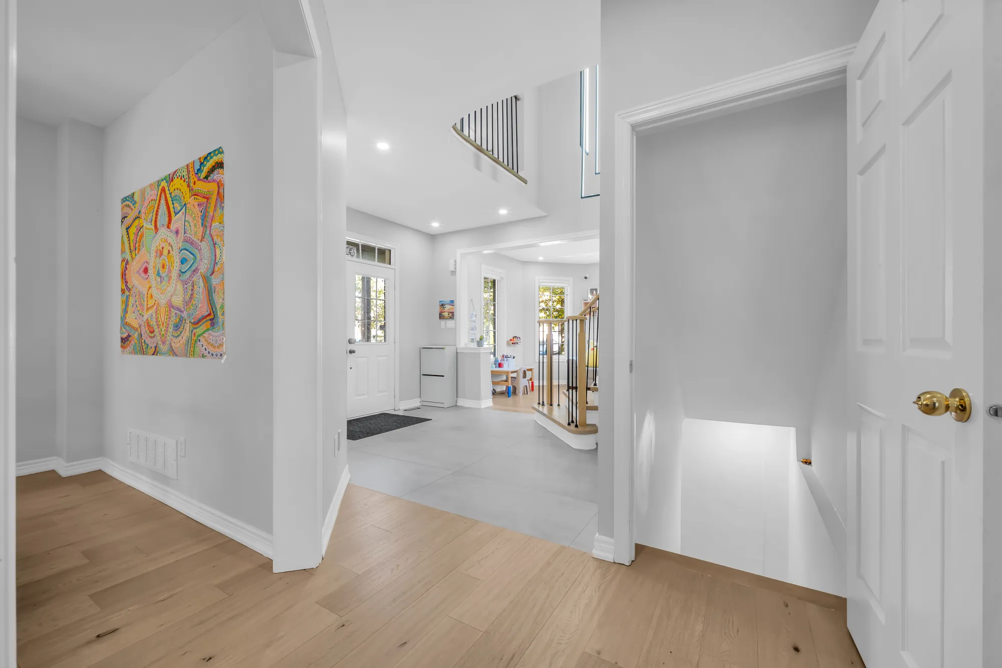 From the dining room toward the foyer and the front doors, a colourful painting at the left, a door open at the right onto a lit stair