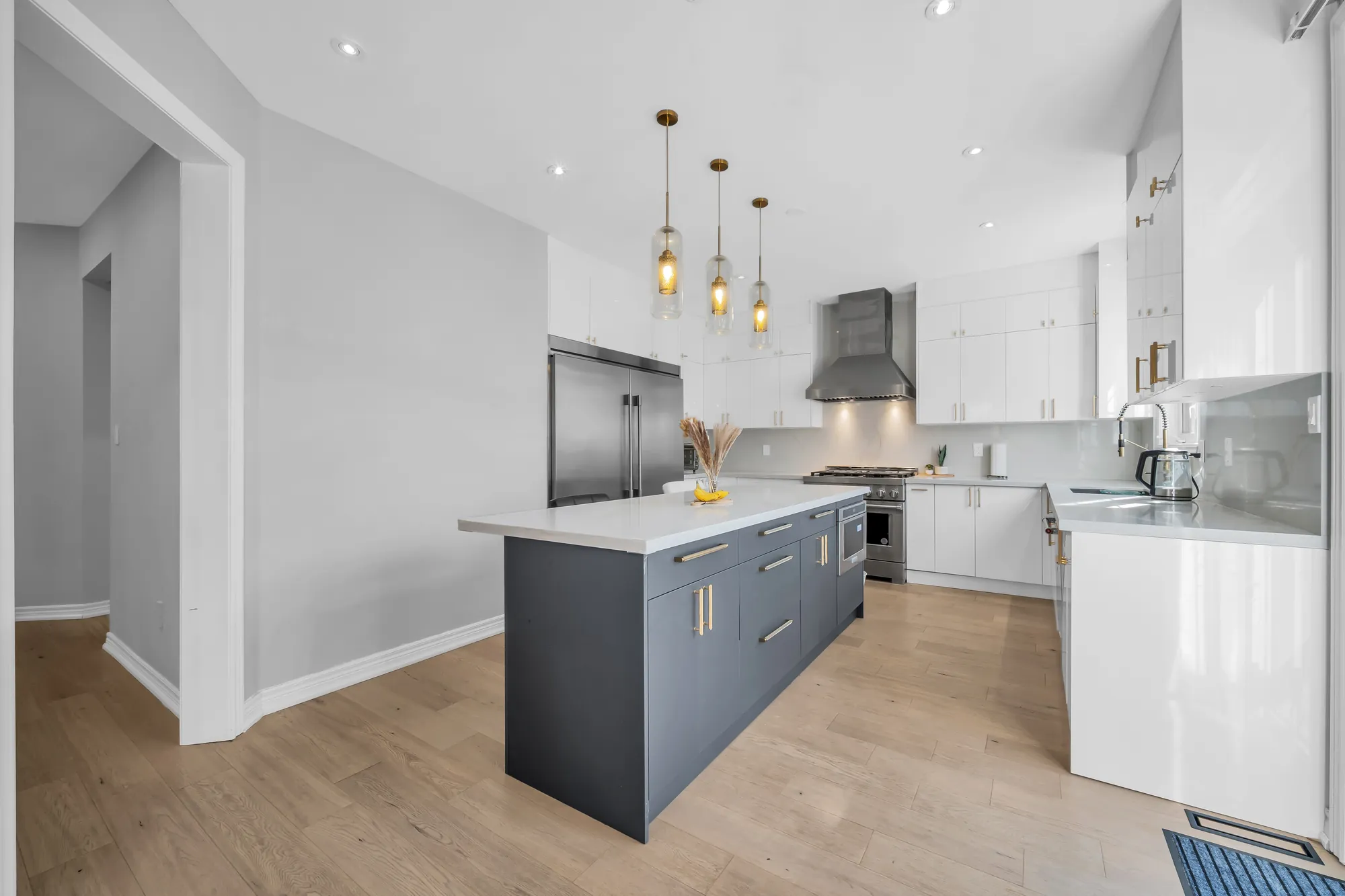 The kitchen: white cabinets to the ceiling, a long grey island with a white quartz top, three glass pendants, a built-in stainless fridge, a range under a stainless hood