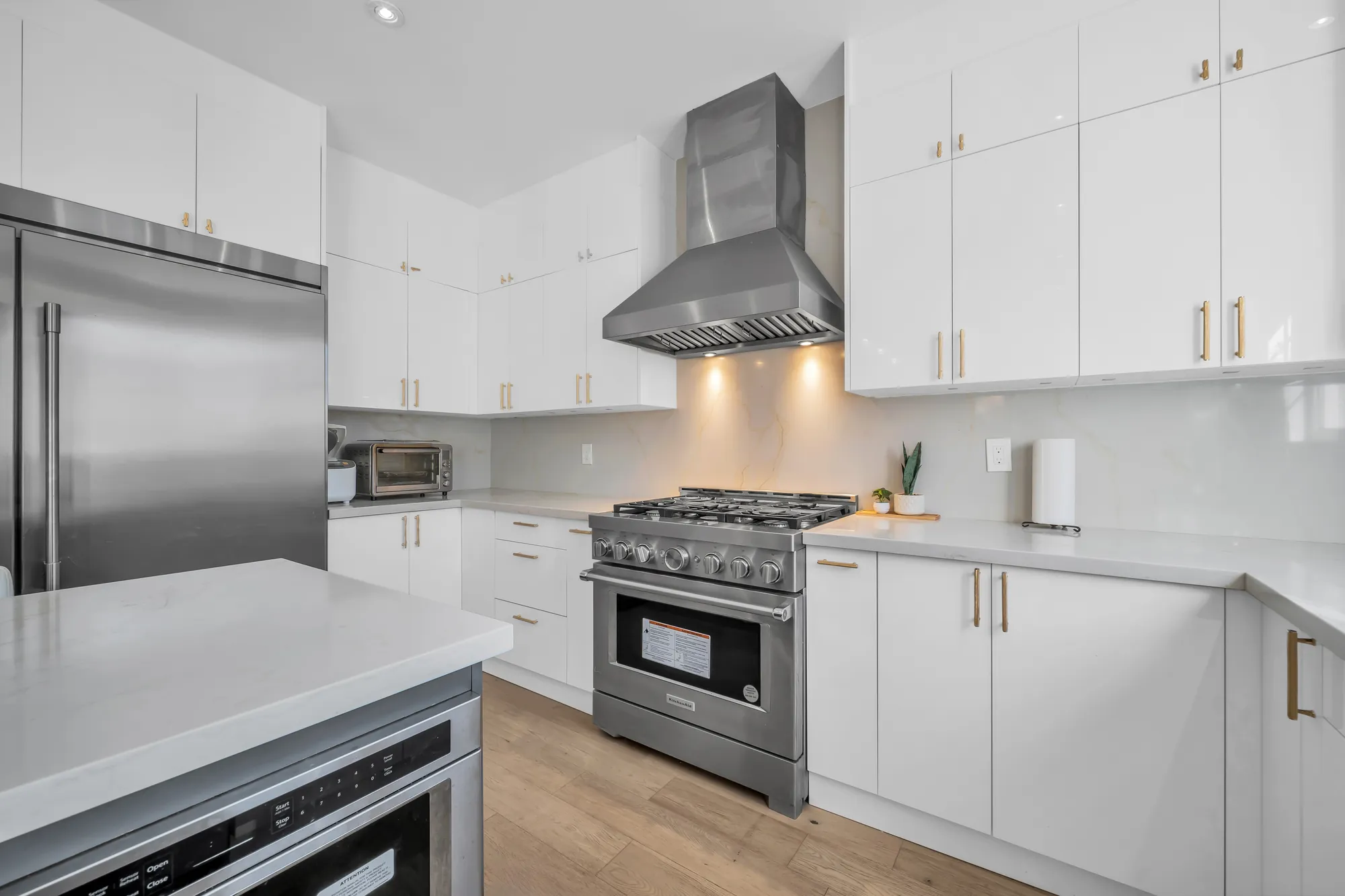 The range wall: a wide stainless gas range under a stainless hood, white cabinets with brass pulls, a white quartz backsplash, the fridge at the left