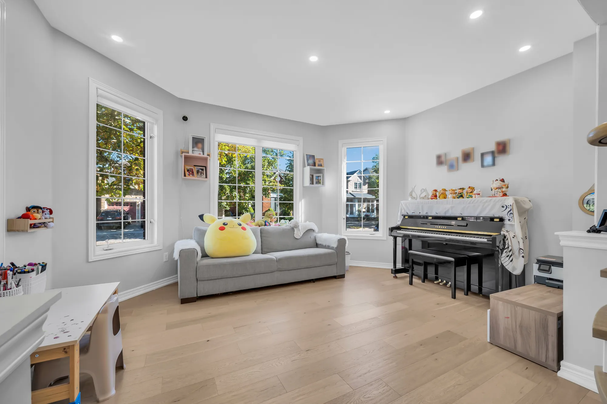 The living room: windows on three sides of a bay, a grey sofa, an upright piano, a child&rsquo;s table and shelves of toys, wood floors