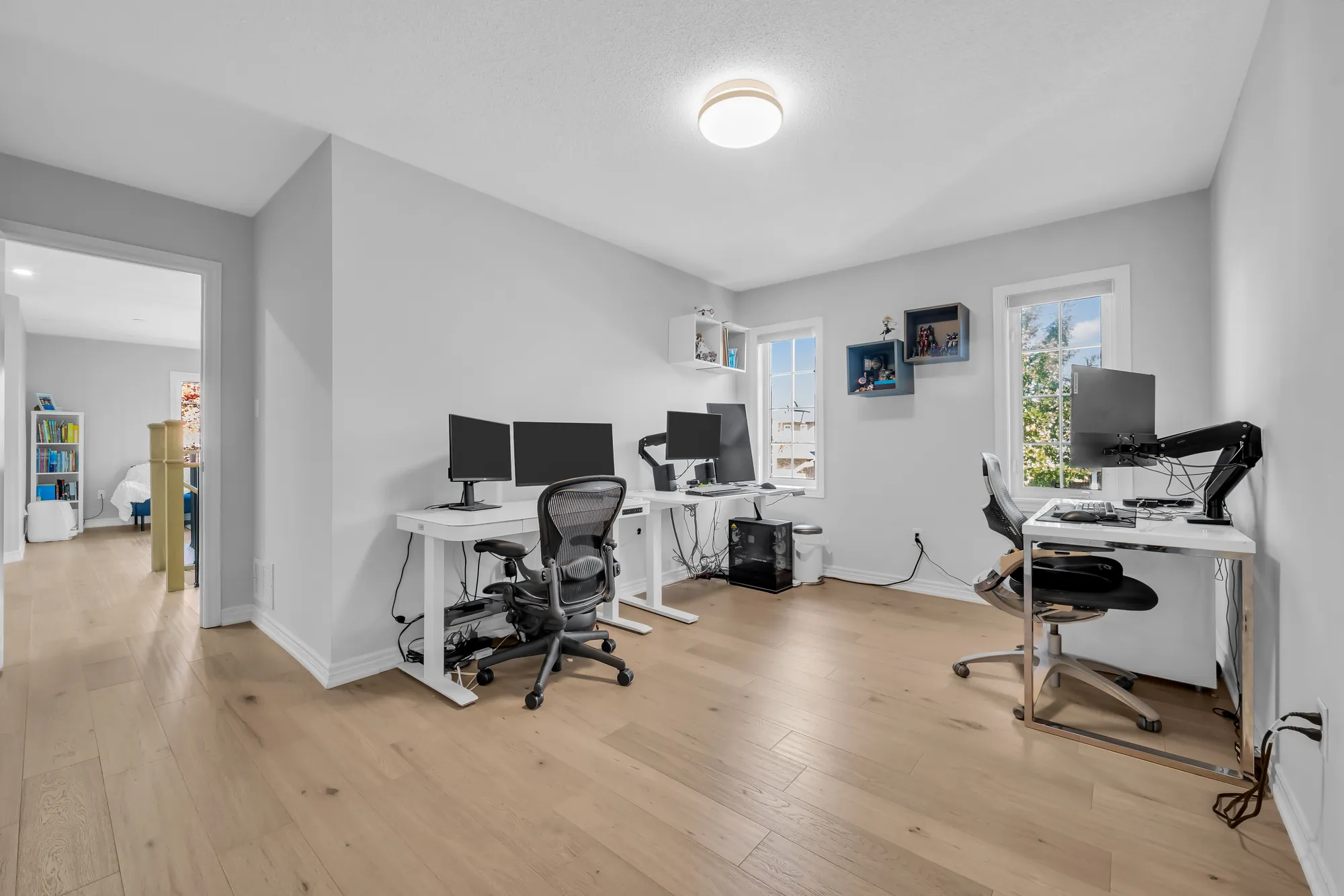 A bedroom used as an office: two desks with monitors and chairs, wood floors, a window at the right