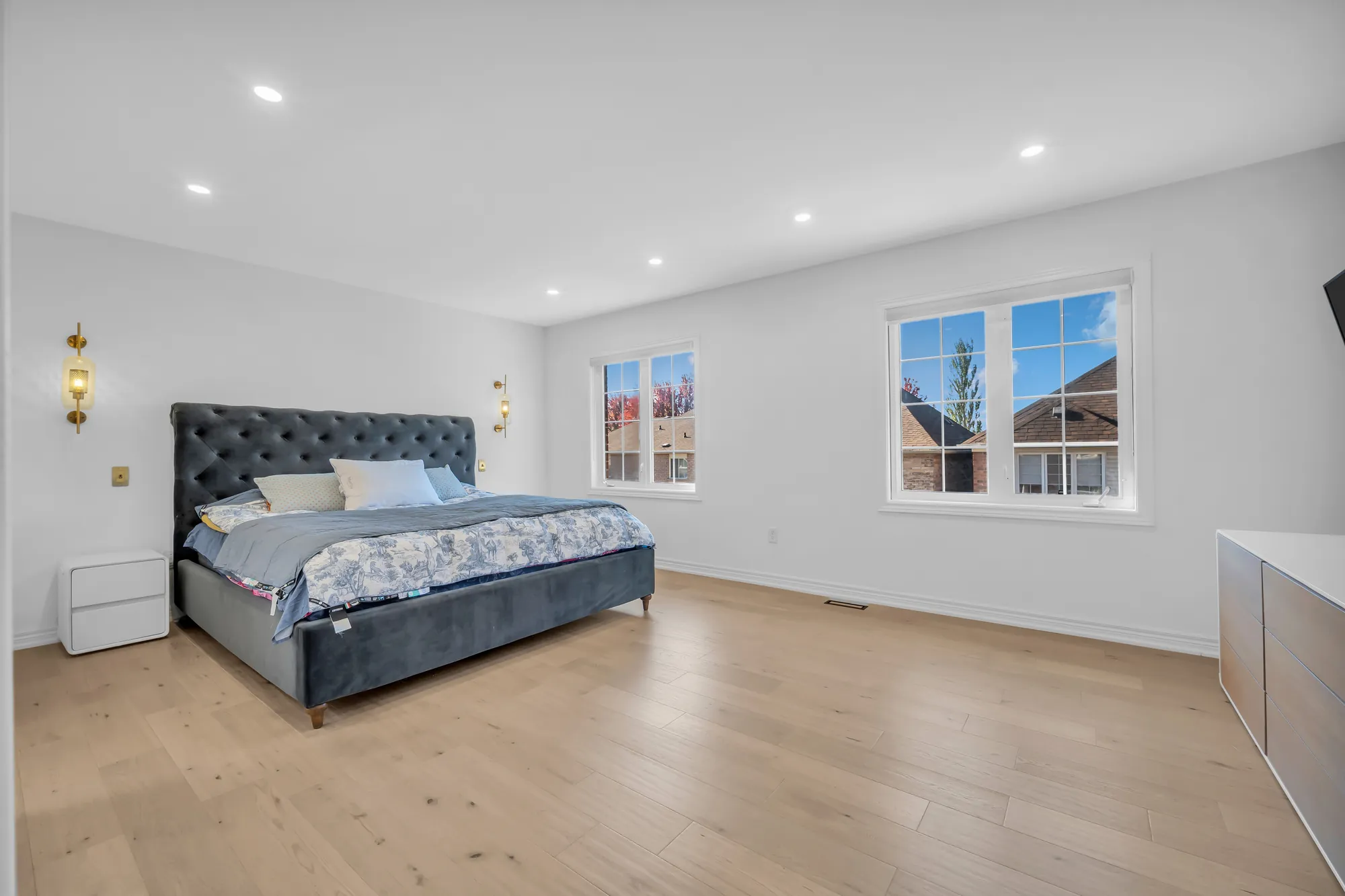 The primary bedroom: a bed with a tufted grey headboard, brass wall lights, two windows, pot lights, wood floors, a dresser at the right