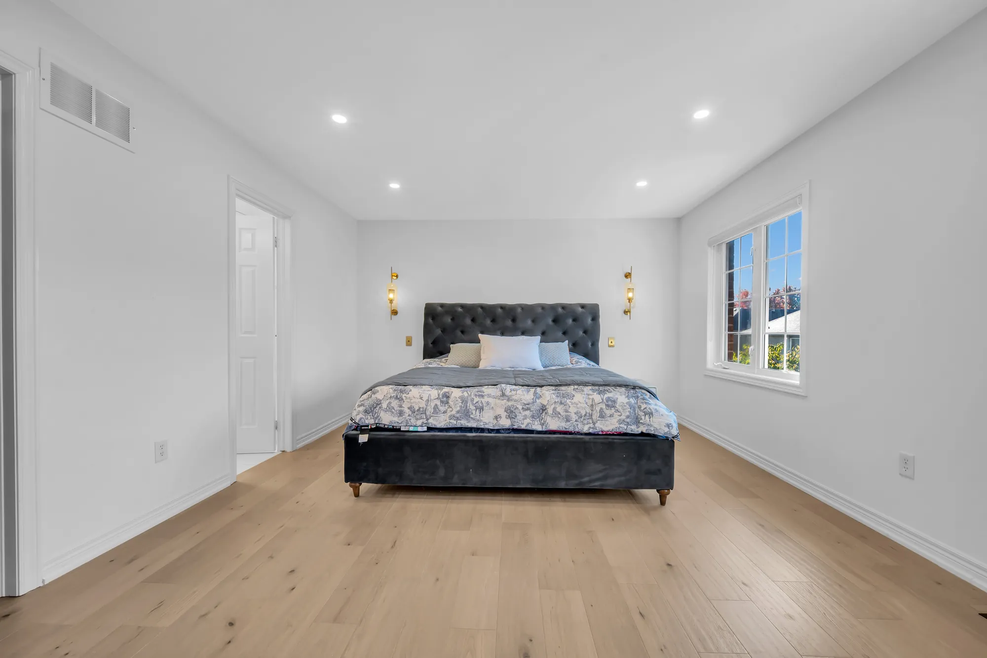 The primary bedroom from the windows: the bed against the far wall, a door at the left, wood floors