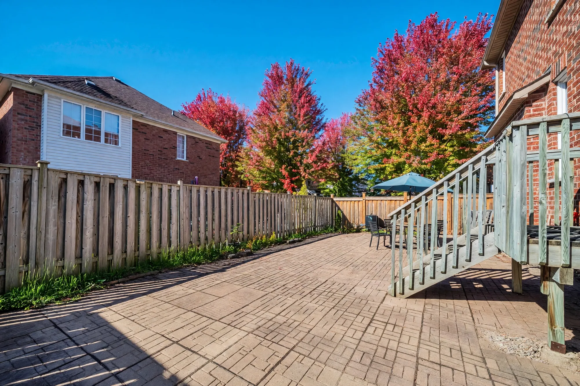 The patio toward the back fence: interlocking stone, the wooden stair down from the deck at the right, a table and chairs under an umbrella in the corner, red maples beyond the fence