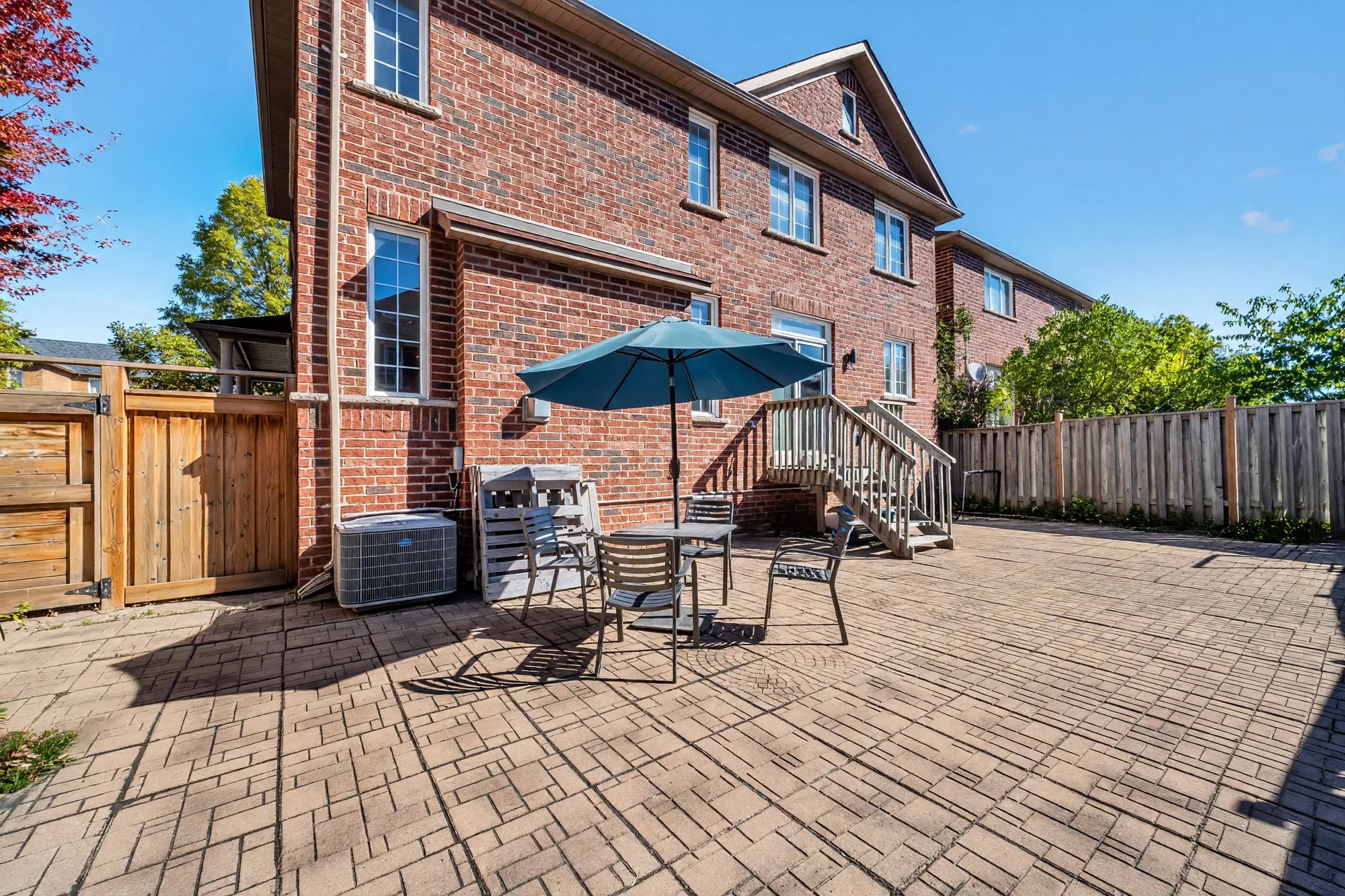 The back of the house over the patio: red brick on two storeys, the deck stair, an air conditioner, a table and chairs under a blue umbrella, a gate in the wooden fence