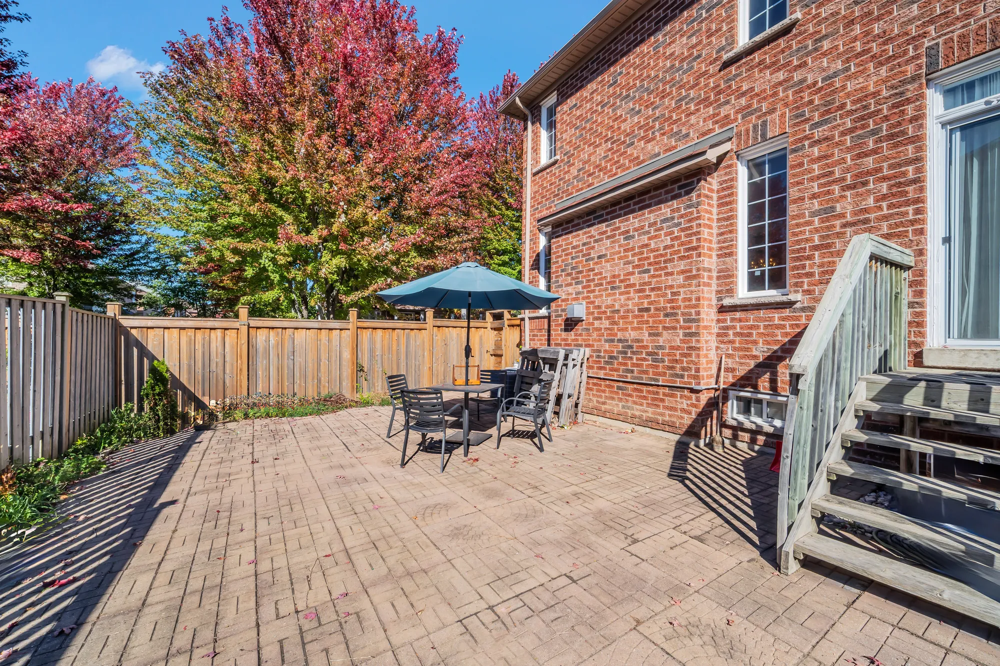 The back yard: an interlocking stone patio, a table and chairs under a blue umbrella, a wooden fence, the back of the house and the stair from the deck at the right, maples beyond